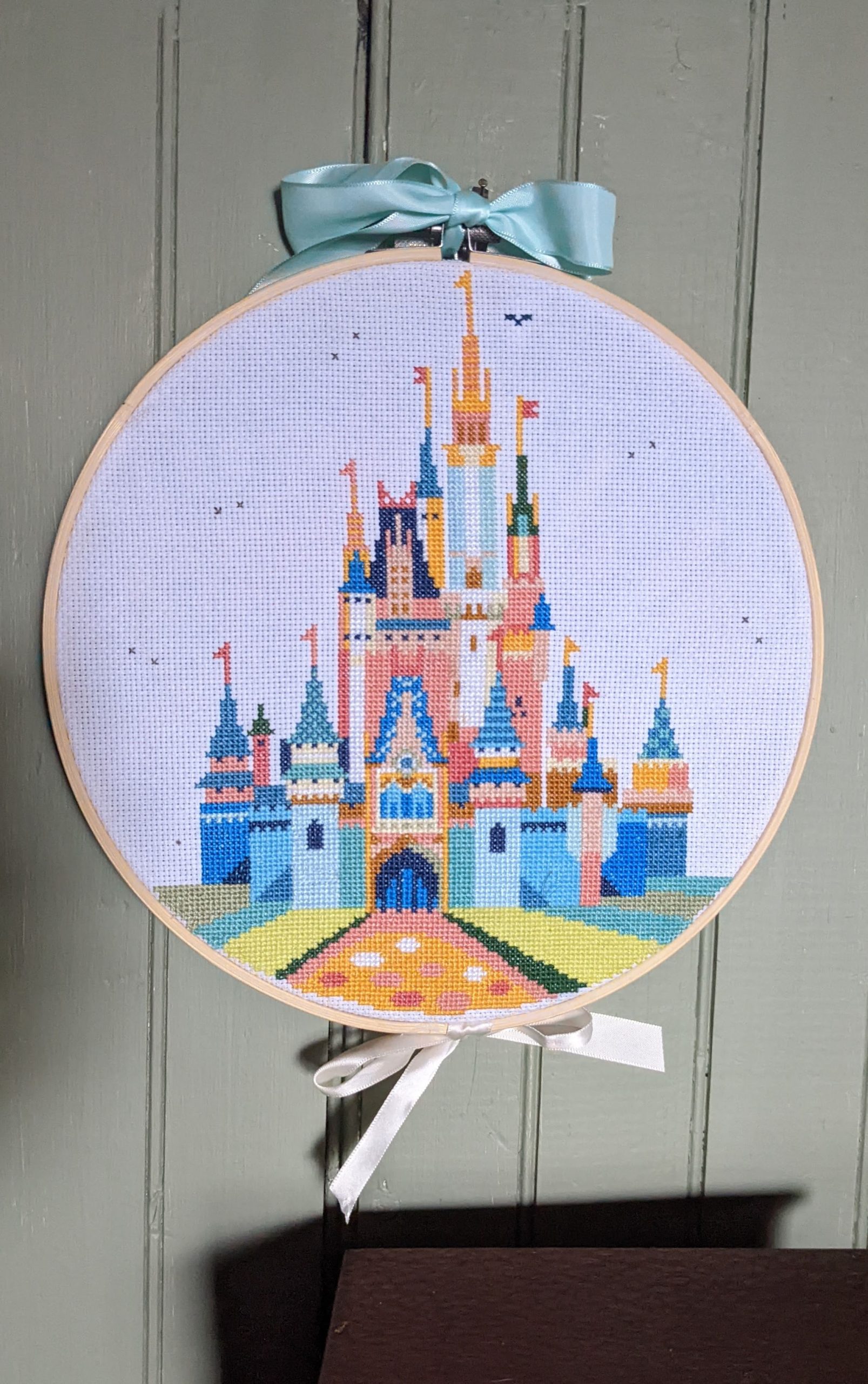Image of a colorful cross stitch of the disney castle on a hoop with bows on a green headboard wall